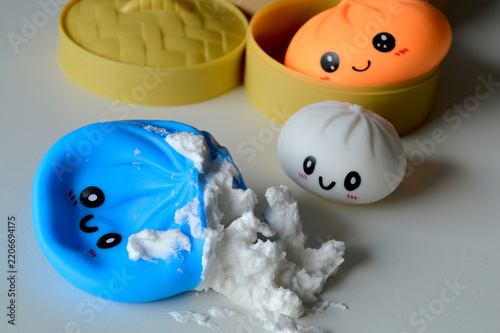 Obraz na plátně Adorable kawaii squishy dumpling toys with smiling faces sit beside a bamboo steamer basket, one blue character split open revealing fluffy white stuffing on a bright surface