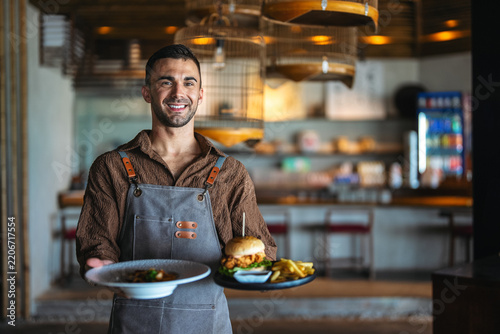 Friendly male waiter in apron serving delicious burger and fries to a guest in cafe