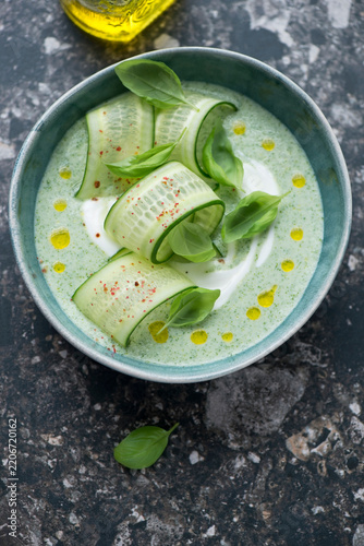 Bowl of chilled cucumber soup topped with cucumber ribbons, yogurt and basil, vertical shot on a dark-brown granite background
