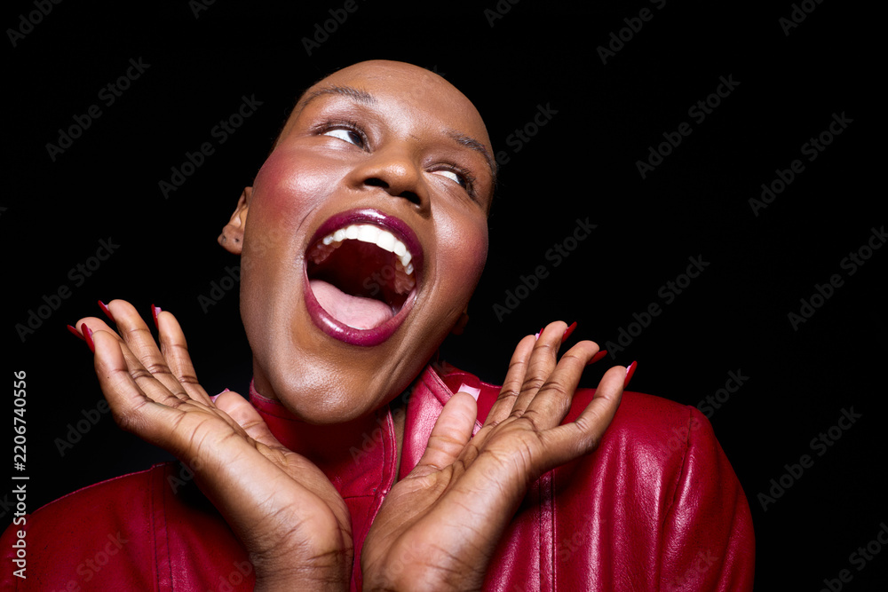 Obraz premium Joyful dark skinned African woman with bald head wearing red jacket laughs with open mouth and hands near face on black background. Concept for happiness, confidence, and beauty.