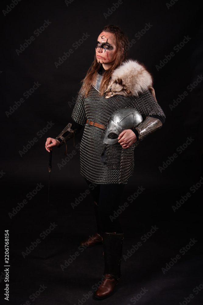 Obraz premium A young Viking woman in fur-trimmed chainmail and war paint, holding a sword, poses against a dark studio background.