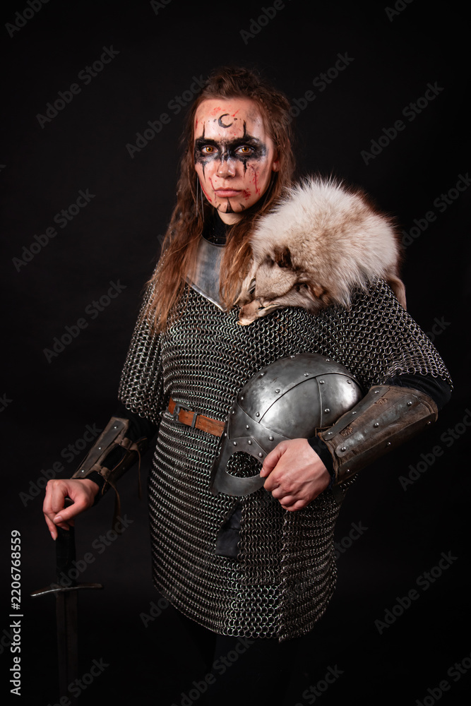 Obraz premium A young Viking woman in fur-trimmed chainmail and war paint, holding a sword, poses against a dark studio background.