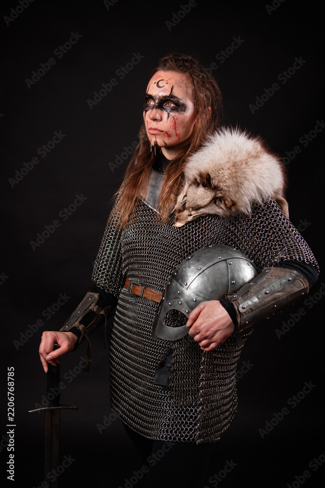 Obraz premium A young Viking woman in fur-trimmed chainmail and war paint, holding a sword, poses against a dark studio background.