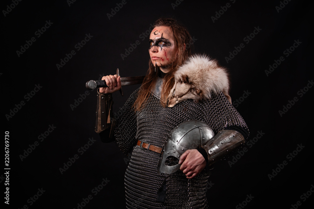 Obraz premium A young Viking woman in fur-trimmed chainmail and war paint, holding a sword, poses against a dark studio background.