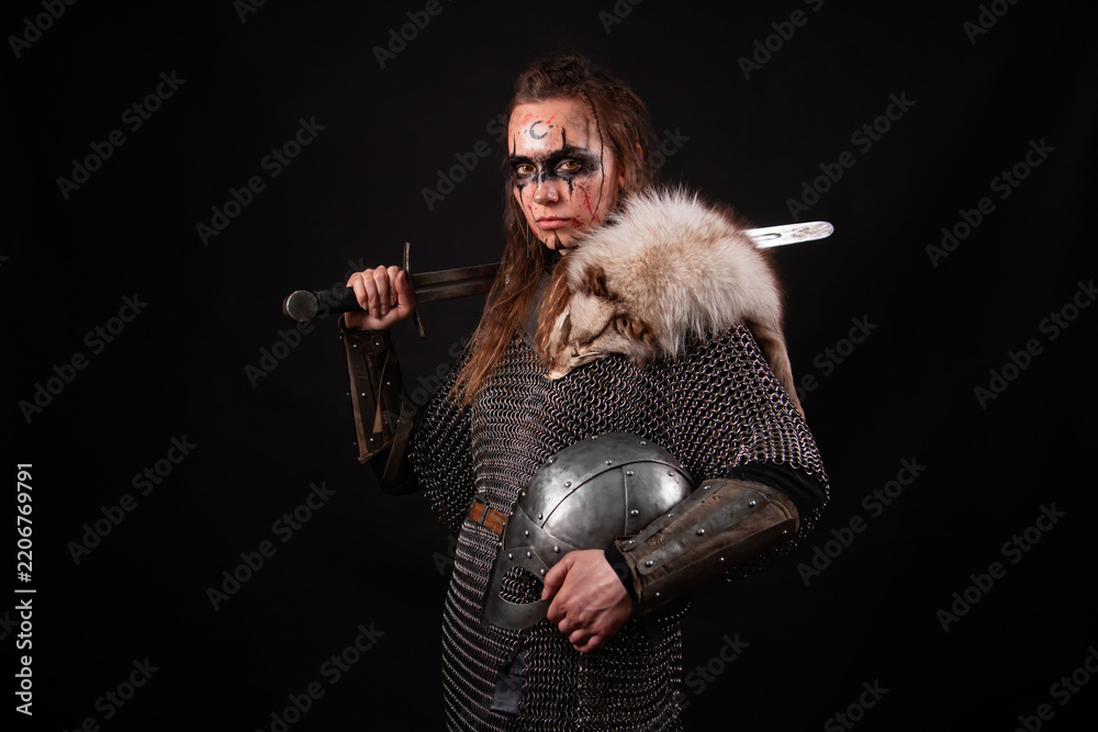 Obraz premium A young Viking woman in fur-trimmed chainmail and war paint, holding a sword, poses against a dark studio background.