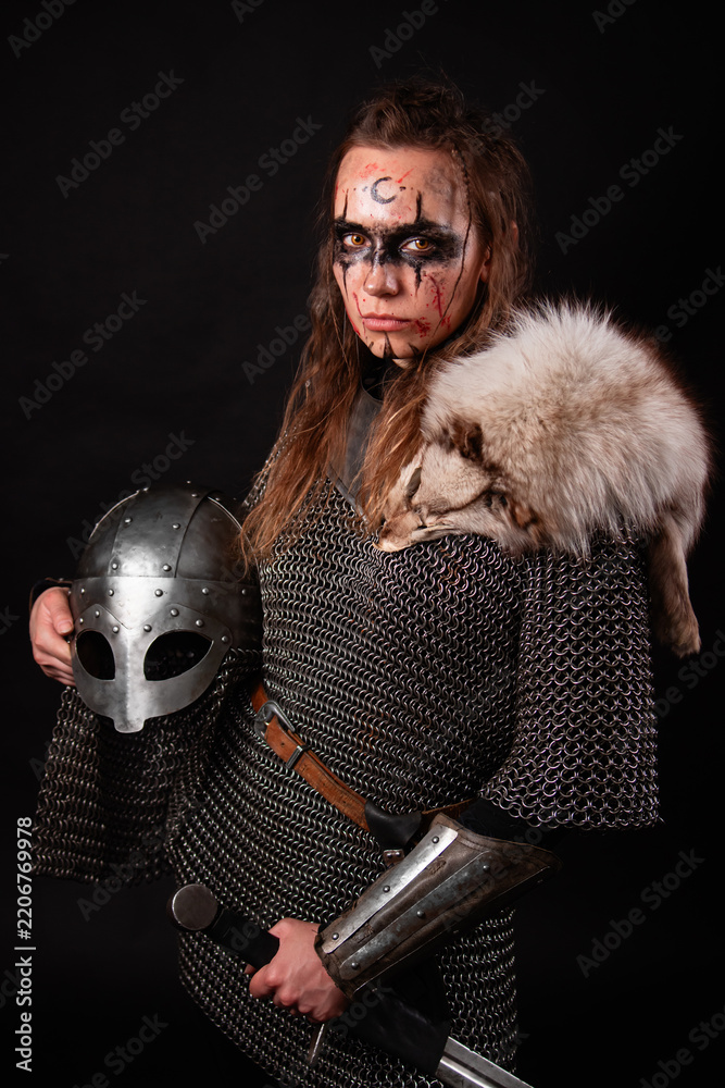 Obraz premium A young Viking woman in fur-trimmed chainmail and war paint, holding a sword, poses against a dark studio background.