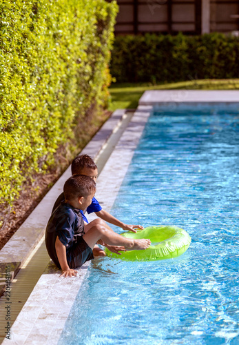Two Cute Asian Brothers Playing Together on the Edge of a Swimming Pool with a Bright Green Inflatable Ring During Summer Vacation