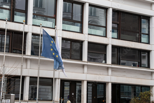 European Union flag flying on a pole among other flagpoles in front of a modern white office building with grid windows and blinds, concept of EU institutions, European politics and international coo