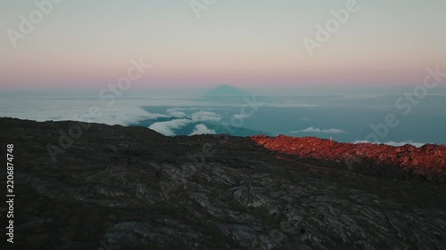 Breathtaking view from the summit of Mount Pico, Azores, looking across a vast sea of clouds towards a distant island, with the volcano's dramatic shadow appearing on the horizon at sunset.