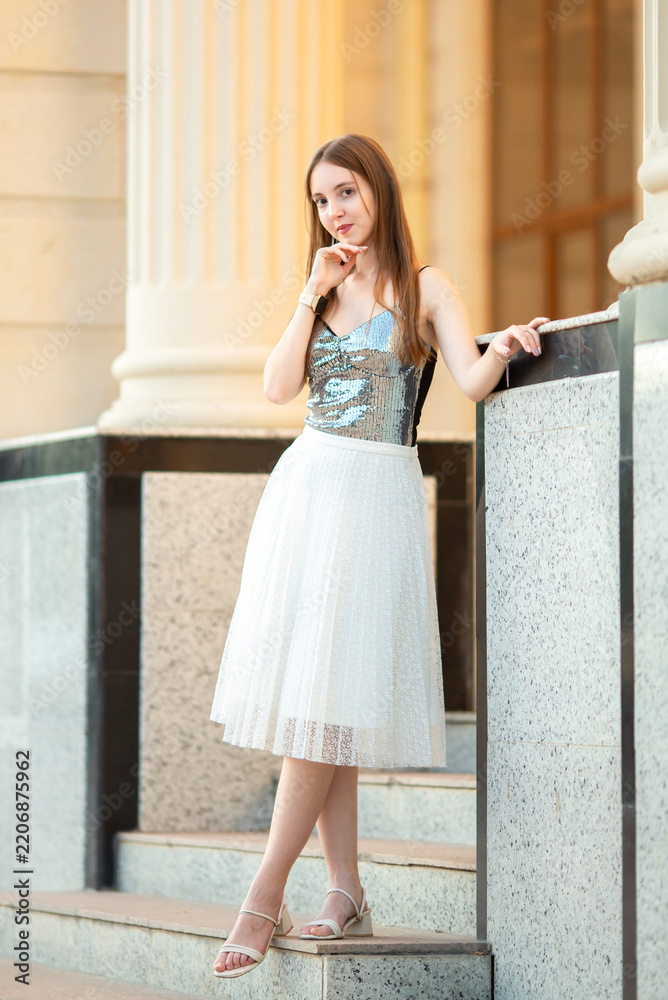 Fototapeta premium An elegant young woman in a stylish outfit poses on the stairs of a classic old building with columns. A fashion portrait, beauty, luxurious lifestyle, confidence, and urban elegance.