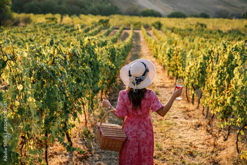 Obraz premium Woman with straw hat holding wine glass and basket walking in vineyard