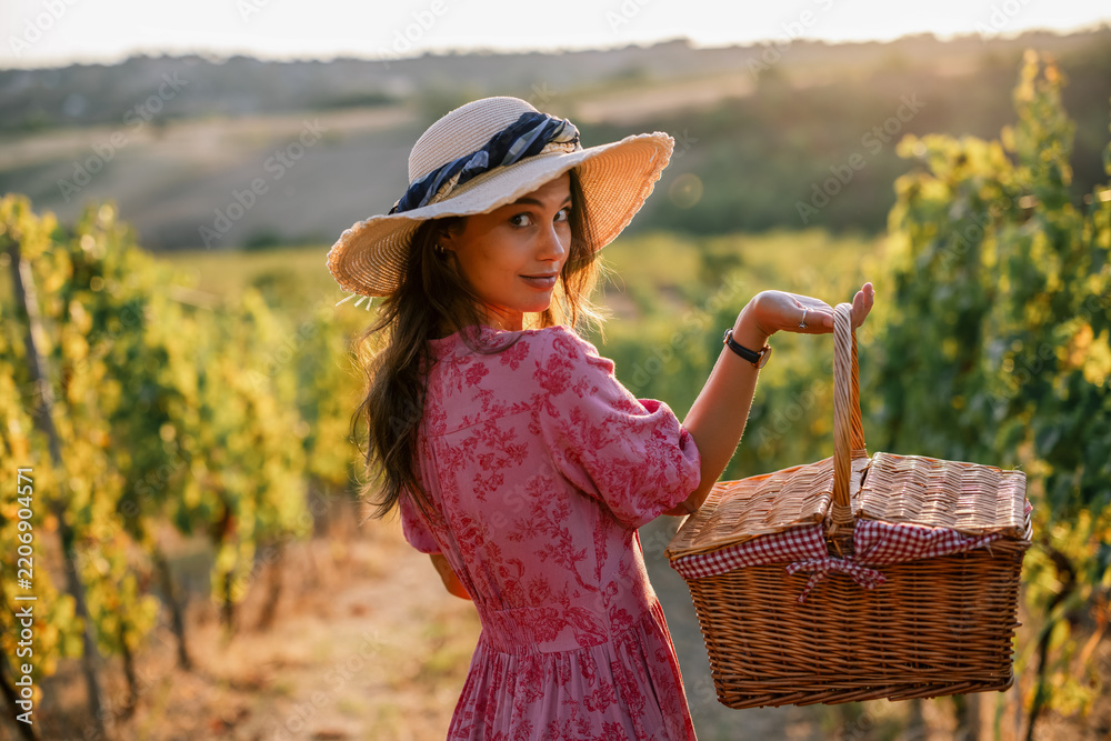 Obraz premium Woman looking over shoulder holding picnic basket in vineyard at sunset