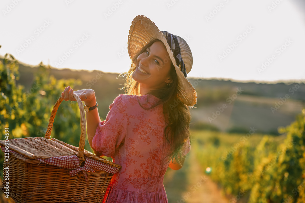 Obraz premium Smiling woman holding wicker basket looking back in sunset vineyard
