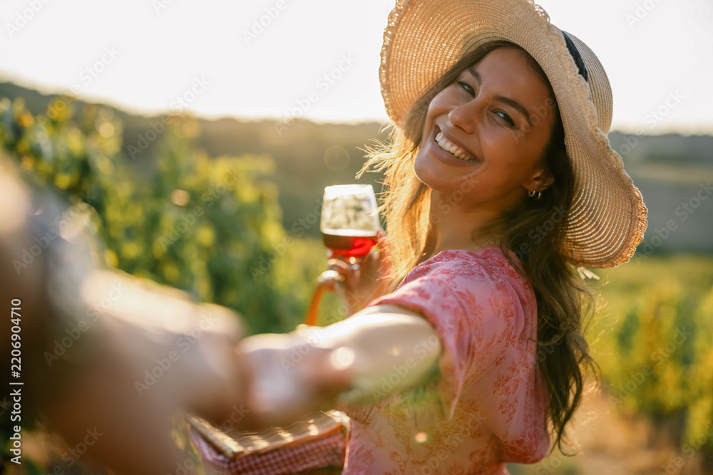 Obraz premium Woman holding hand leading partner through vineyard holding wine glass at sunset
