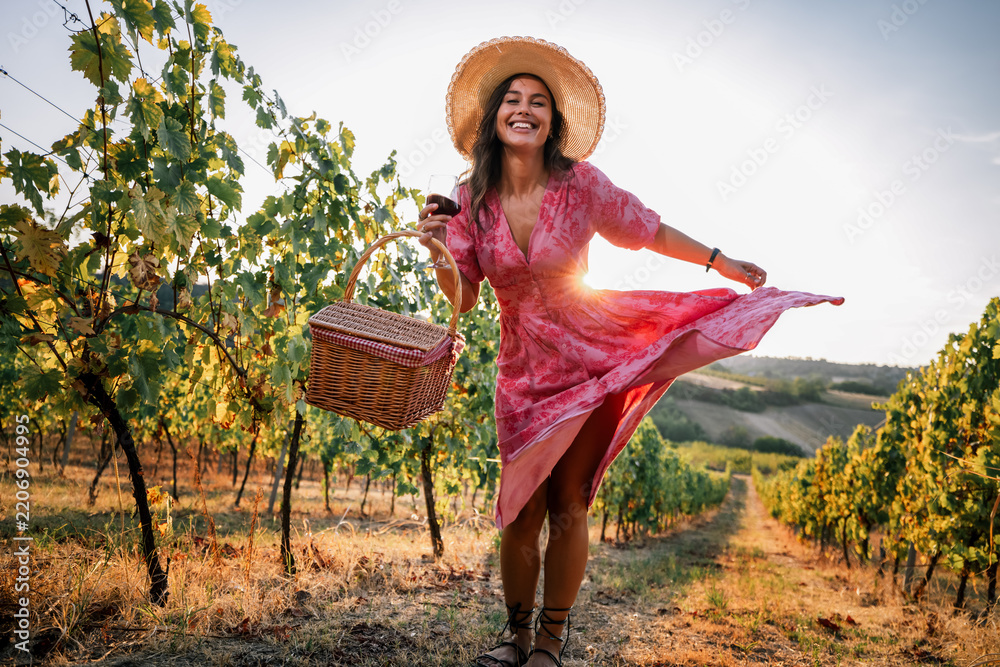 Obraz premium Playful woman twirling pink dress in vineyard holding picnic basket and wine