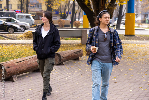 Young man with headphones and a girl in a black hoodie walking along a paved path in a city park with yellow leaves.