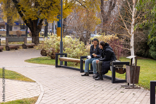 Two young people sitting on a rustic bench in a park with yellow autumn trees, having lunch and talking.
