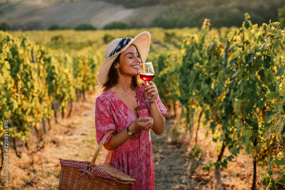 Obraz premium Woman in straw hat smelling glass of red wine in vineyard at sunset