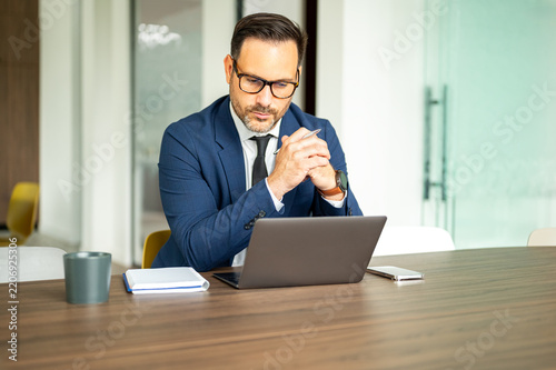 Business professional at a desk, focused on a laptop while reviewing financial figures and legal documents, contemplating strategy and critical decisions in an office setting