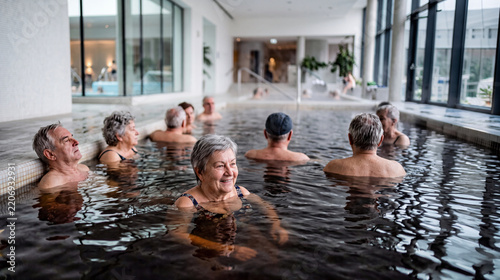 Smiling senior woman relaxing in an indoor thermal bath pool with a group of elderly men and women. Bright modern spa interior, bathing, wellness, leisure and healthy lifestyle in retirement.