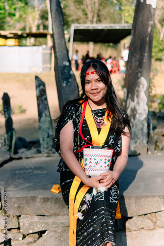 Indonesian woman in Bajawa Ngada traditional attire posing with pressed palms at megalithic site, expressing hospitality and cultural heritage.
