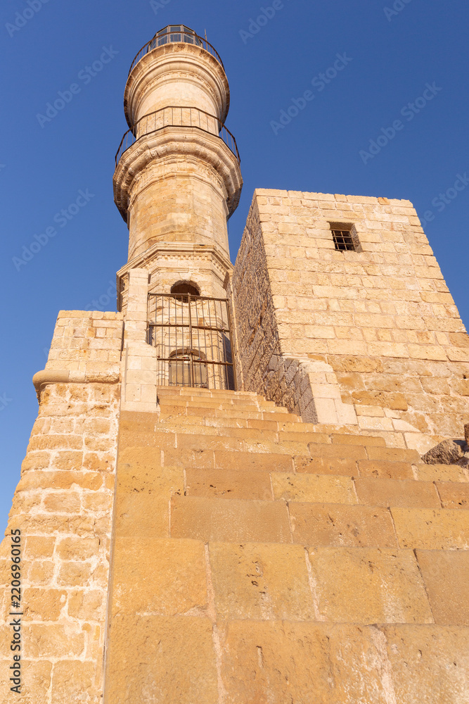 Fototapeta premium Historic Venetian lighthouse and stone harbor wall illuminated by warm sunrise light in Chania, Crete, Greece.