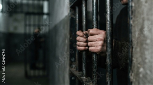 Hands grip rusted bars with visible wear. Fingers show strain, suggesting confinement. Blurry figure stands in dim, shadowy background. Damp concrete walls emphasize bleakness