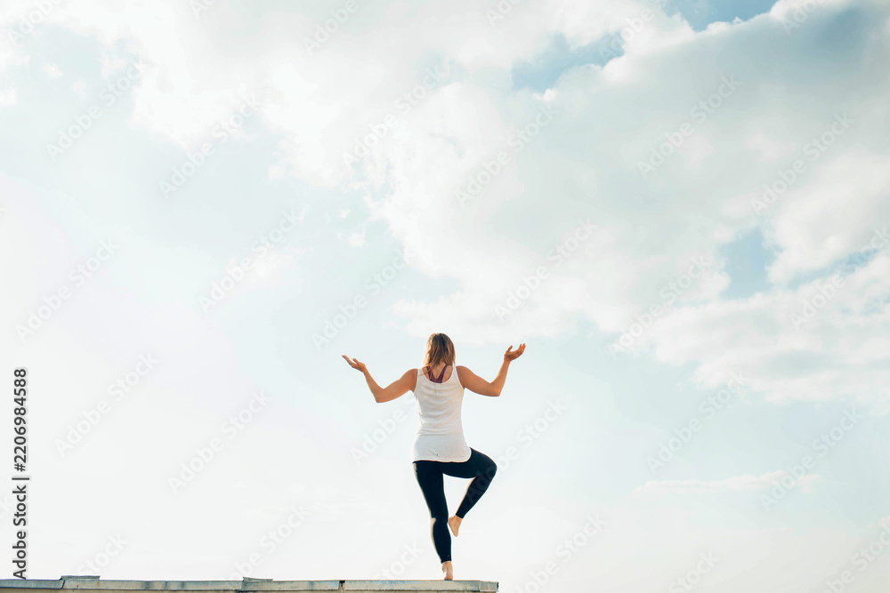 Obraz premium Young woman practices yoga outside. Blonde girl turned back standing on one leg, second raised bent at knee. Hands on sides bent at elbows and turned up. Sky and clouds on background.