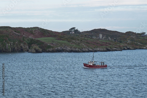 A red fishing boat travels across blue water near rocky cliffs and green hills under a cloudy sky.