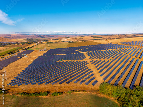 Photovoltaic field with diagonal panel rows, aerial view. Blue arrays occupy dry grass above a cultivated foreground, with hills along the horizon.