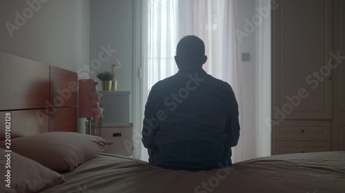 Rear view of a lonely, depressed man sitting on the edge of his bed in a dark bedroom, contemplating his problems and feeling anxious, representing mental health issues and sadness