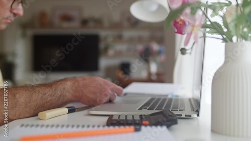 Frustrated man managing finances at his home office desk, checking bills and receipts while calculating expenses on a laptop, feeling the pressure of budget problems and economic crisis