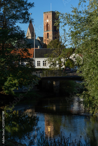Vertical composition showing the prominent stone tower of Ribe Cathedral rising behind a peaceful river channel, arched bridge, and lush green trees