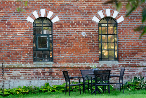 Symmetric view of a historic red brick facade featuring decorative arched windows and a cozy black patio table and chairs set on a green lawn.