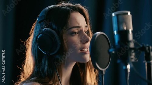 Singer Recording in Studio: A young woman with headphones on intensely focuses as she sings into a professional microphone in a dimly lit recording studio, her passion evident in her expression.