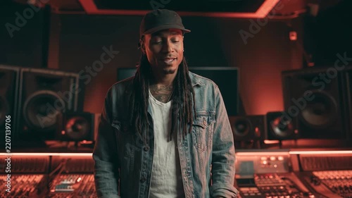 Producer in Sound Studio: A thoughtful man with dreadlocks, wearing a cap and denim jacket, stands amidst the professional audio equipment of a recording studio.