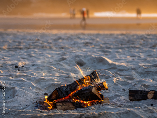 Small beach bonfire burning in the sand at Cannon Beach, Oregon, with warm sunset light, shallow depth of field, and blurred people walking near the shoreline in the background