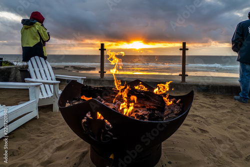A person relaxing beside a glowing fire pit on an oceanfront terrace at sunset, with dramatic clouds, golden light, and Pacific waves along the Oregon Coast