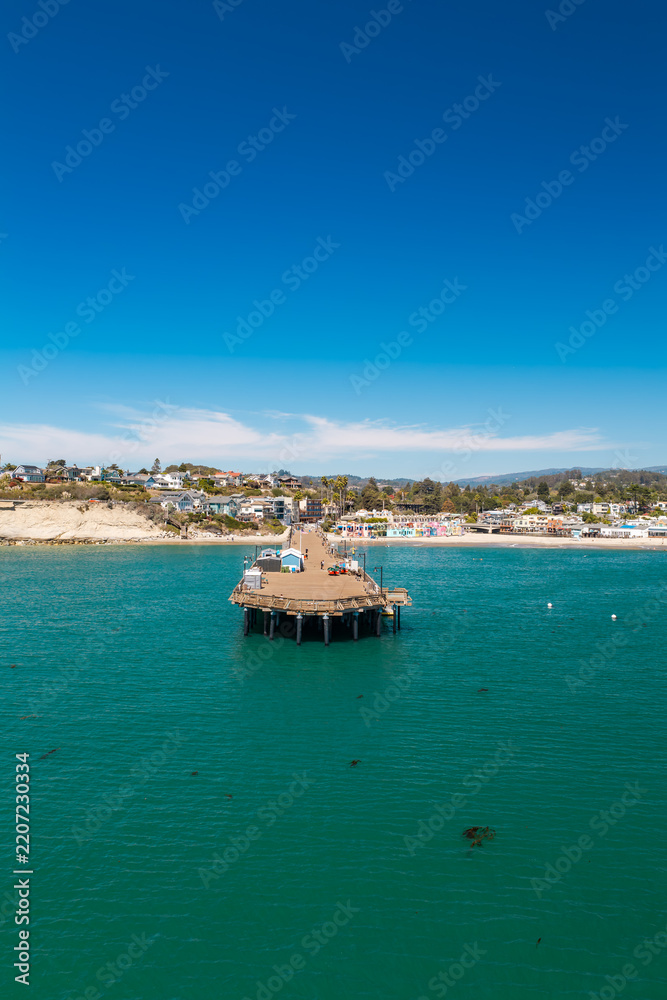 Fototapeta premium Aerial view captures Capitola Wharf extending into the ocean towards the sandy beach and coastal town buildings in Capitola, California, under a clear sky.