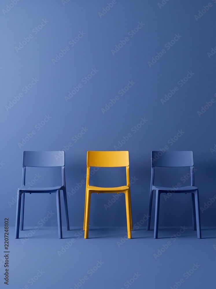 Obraz premium Single yellow plastic chair standing out between two blue chairs against blue wall.
