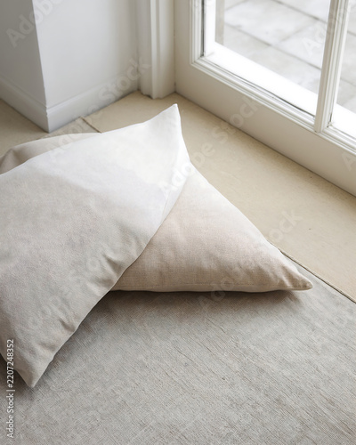 Two rectangular linen pillows in shades of beige and off white resting on a light colored floor surface near a large glass window with white frame