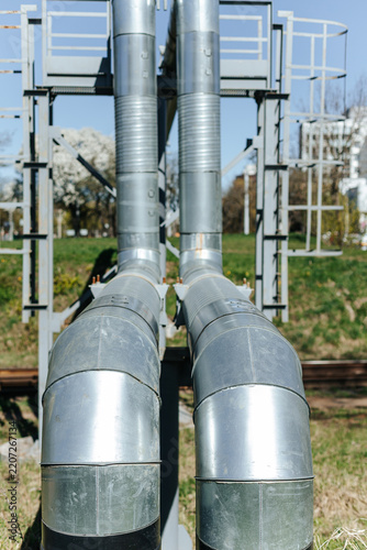 Insulated Industrial Pipes Curve Along Steel Support Framework. Outdoor Utility Infrastructure Stands Near Railroad Tracks
