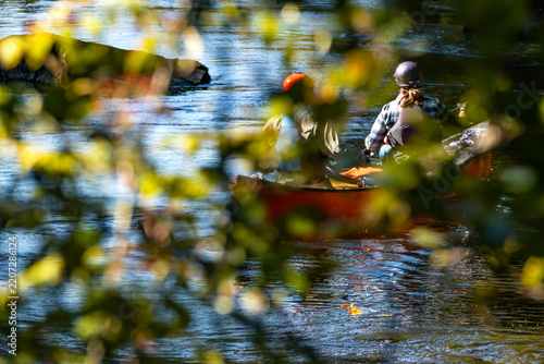 A View Through the Blurry Leaves of a Tree of Two People in Helmets in a Red Canoe Heading Towards a Rapid