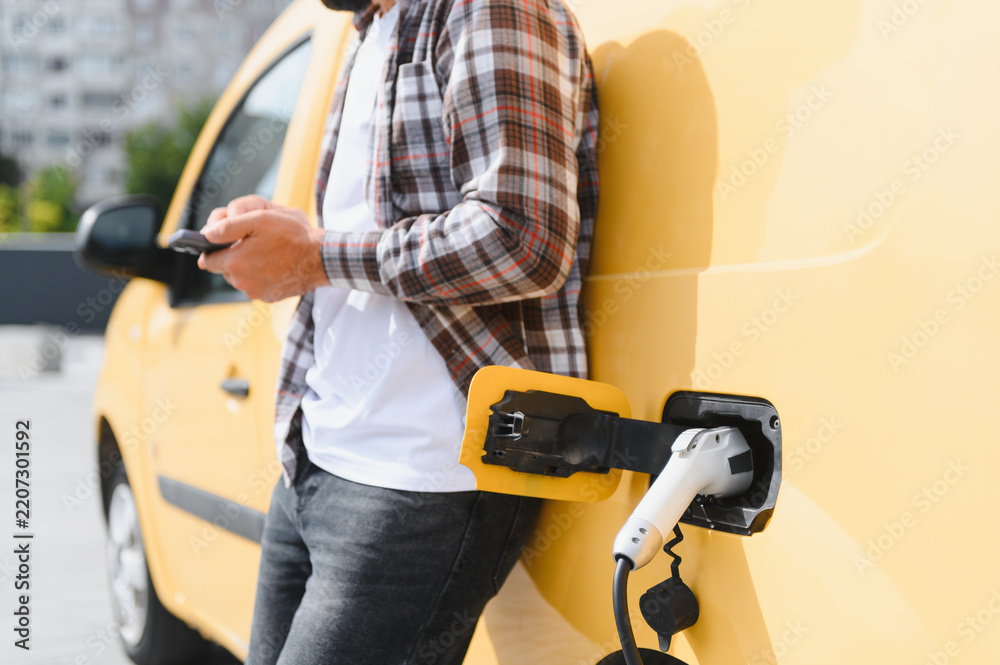 Fototapeta premium Deliveryman using smartphone while charging electric van