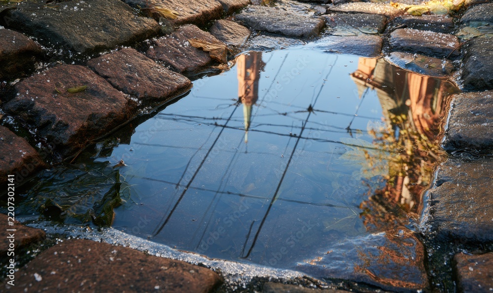 Fototapeta premium Reflection of church tower and trees in shallow puddle among cobblestones