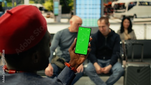 Fotografía Black man holds a vertical chroma key phone in a busy bus station while passengers wait for an intercity coach; the mockup screen appears in a crowded terminal
