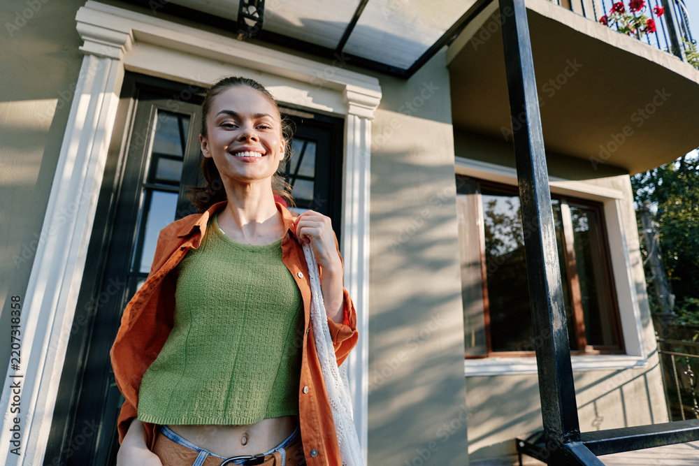 Obraz premium Smiling young woman in a green knitted top and orange shirt walks near a house with a white bag. Sunny day, casual style. Concept of lifestyle and happiness.