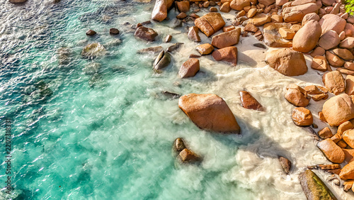 Aerial drone view of the tropical beach on Praslin Island, Seychelles, featuring turquoise waters, white sand, lush tropical vegetation, and granite rock formations along the Indian Ocean coastline.	

