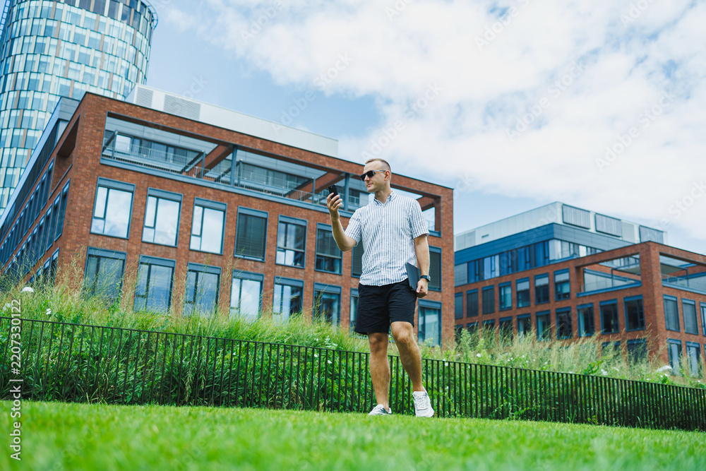 Fototapeta premium Man walks on grass while holding a smartphone near modern buildings during daytime