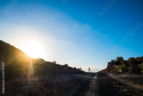 Fotografie Lone runner sprinting along a rocky desert trail at sunrise, captured as a dramatic silhouette against a clear blue sky